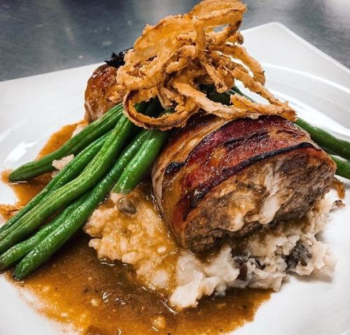 A plated dish featuring meatloaf wrapped in bacon, topped with crispy onions, served with green beans and mashed potatoes.