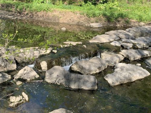 A small waterfall cascades over rocks into a calm stream, surrounded by lush greenery.