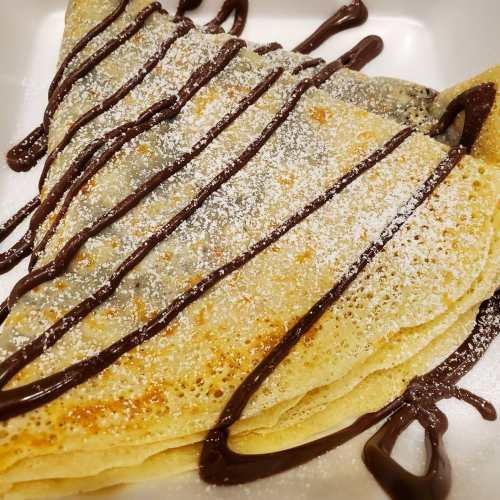 A stack of crepes drizzled with chocolate sauce and dusted with powdered sugar on a white plate.