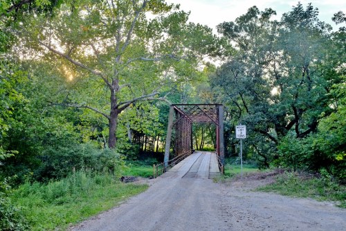A rustic metal bridge spans a dirt road, surrounded by lush green trees and soft sunlight filtering through the foliage.