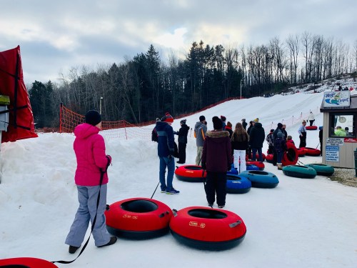 A group of people waiting in line with snow tubes at a winter tubing hill, surrounded by snow and trees.