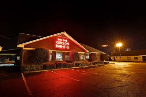 A barbecue restaurant at night, featuring a red neon sign that reads "Big Bob Gibson Bar-B-Q" and illuminated parking lot.