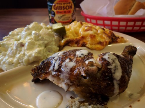 A plate featuring grilled chicken with sauce, macaroni and cheese, potato salad, and a drink in the background.