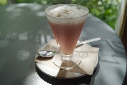 A glass of pink beverage with foam, served on a saucer with a spoon, set on a table with soft natural light.