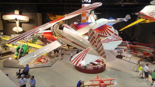 A vintage biplane with red and white stripes is displayed in an aviation museum, surrounded by various aircraft.