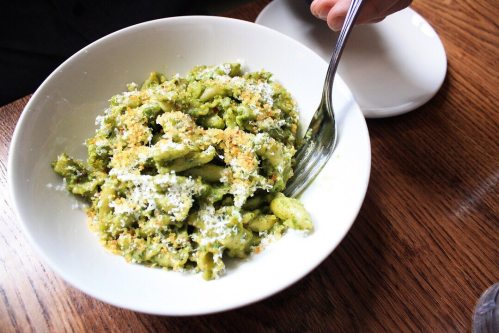 A bowl of green pasta topped with cheese and breadcrumbs, with a fork resting on the side.