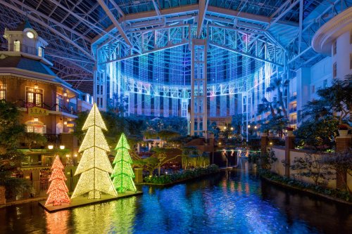 Colorful illuminated Christmas trees by a river inside a large atrium, with vibrant lights and greenery in the background.
