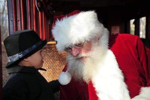 A child in a hat talks to Santa Claus, who is wearing a red suit and white beard, inside a train car.