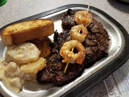 A plate featuring grilled steak, shrimp skewers, cheesy potatoes, and a slice of toasted bread.