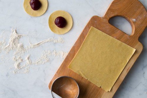 A wooden cutting board with pasta dough, two filled circles, and a round cutter on a marble surface.
