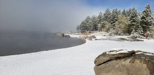 A snowy shoreline with large rocks, surrounded by evergreen trees and fog over the water.