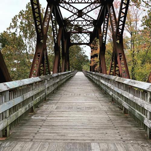 A wooden bridge with metal supports, surrounded by trees in autumn colors, leading into the distance.