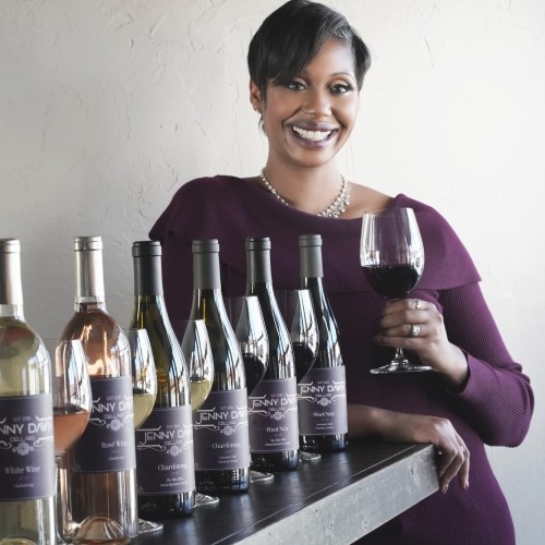 A woman in a purple dress smiles while holding a glass of red wine, surrounded by various wine bottles on a table.
