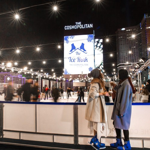 Two people in winter attire stand by an ice rink at night, with a large sign for The Cosmopolitan in the background.