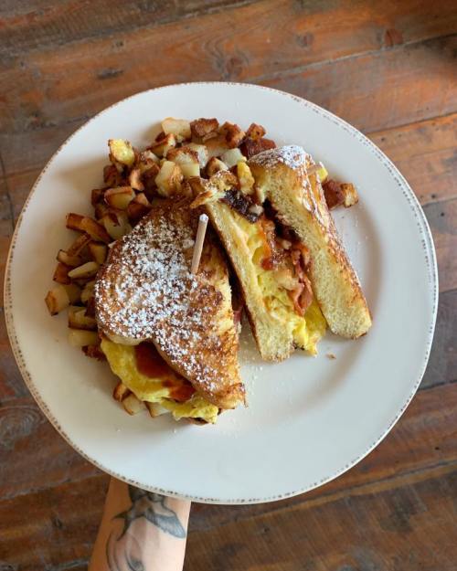 A plate of French toast stuffed with eggs and bacon, served with crispy potato cubes, held by a hand.