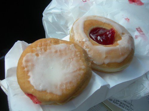 Two donuts on a white paper wrapper: one with a glazed top and jelly filling, the other with a plain icing.