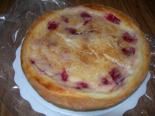A round cheesecake with a golden crust and swirls of red fruit on top, placed on a white cake board.