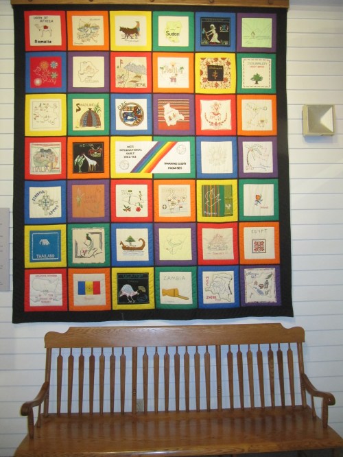 A colorful quilt with various embroidered squares hangs on a wall above a wooden bench.