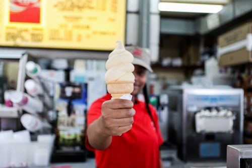A person in a red shirt holds up a soft-serve ice cream cone in a shop, with a menu in the background.