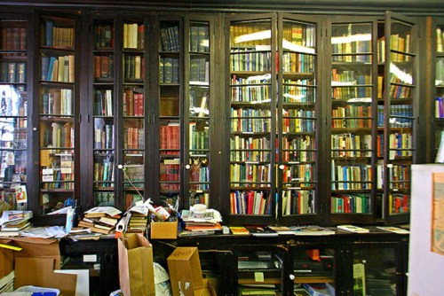 A cluttered library with tall bookshelves filled with colorful books and scattered papers and boxes on the floor.