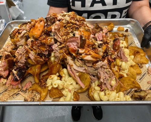 A tray piled high with pulled pork, macaroni, and chips, topped with barbecue sauce, held by a person in a black shirt.