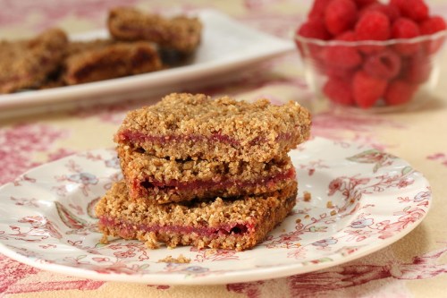 Three stacked raspberry bars on a floral plate, with a bowl of fresh raspberries in the background.