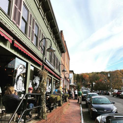 A charming street scene with people dining outside, colorful buildings, and autumn foliage in the background.