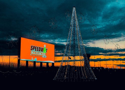 A brightly lit Christmas tree with a billboard displaying "Speedway Christmas" against a sunset sky.