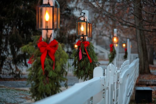 A row of lanterns with warm light and festive wreaths adorned with red bows, beside a white fence in a winter setting.