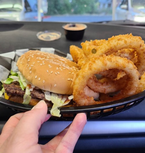 A hand holding a plate with a sesame seed burger and crispy onion rings, set against a car interior backdrop.