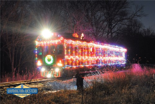 A brightly lit train decorated with colorful holiday lights, surrounded by trees at night. Wilmington & Western Railroad logo visible.