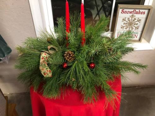 A festive centerpiece with pine branches, red candles, a pinecone, and ornaments on a red tablecloth.