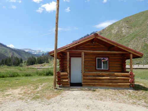 A rustic log cabin with a red roof, surrounded by green fields and mountains under a blue sky.