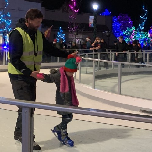 A man in a safety vest assists a child on ice skates at a festive outdoor rink with colorful lights in the background.
