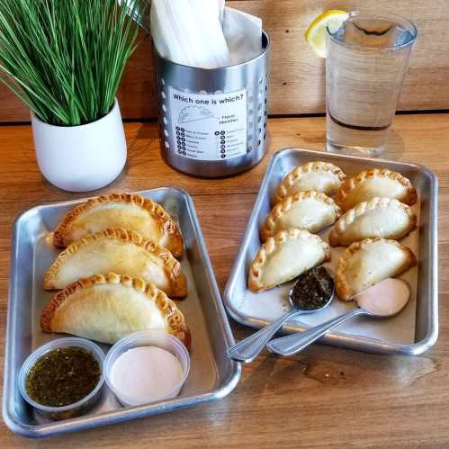Two trays of golden-brown empanadas with dipping sauces, a glass of water, and a plant in the background.
