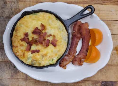 A cast iron skillet with a cheesy egg dish topped with bacon, served alongside crispy bacon and orange slices.