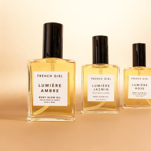 Three bottles of French Girl body glow oil in amber, jasmine, and rose scents, displayed against a soft beige background.