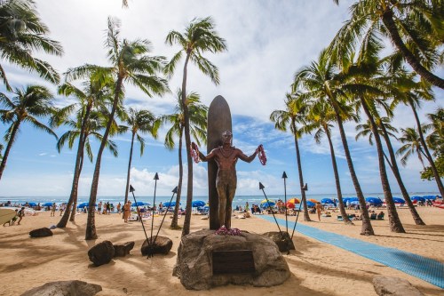 Statue of a surfer with leis, surrounded by palm trees and beachgoers under a blue sky.