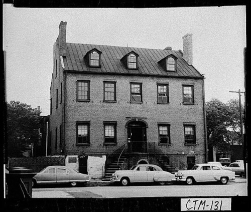 Historic brick building with a steep roof, featuring multiple windows and parked vintage cars in front.