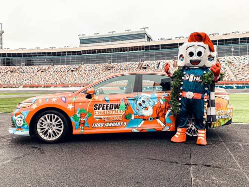 A festive car decorated for a Christmas event, with a mascot in a Santa hat standing beside it at a racetrack.