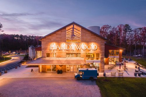 A rustic brewery building with large windows and a sign reading "Hardywood," surrounded by trees at dusk.