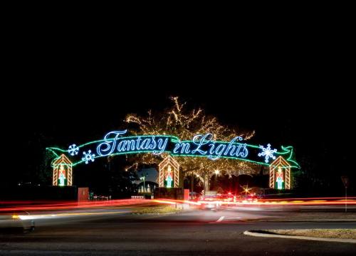 Colorful holiday lights spell "Fantasy in Lights" over a festive entrance, with light trails from passing cars at night.