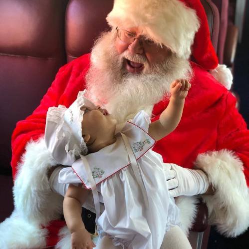 A joyful Santa Claus holds a baby in a white dress, both smiling and sharing a festive moment.