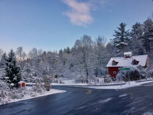 A snowy landscape featuring a red barn and trees, with a clear blue sky and a winding road.