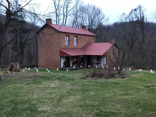 A two-story brick house with a red metal roof, surrounded by trees and a grassy yard, illuminated at dusk.
