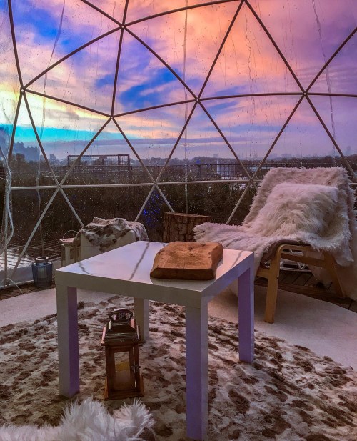 Cozy interior of a geodesic dome with a table, fur rugs, and a colorful sunset view through the transparent walls.