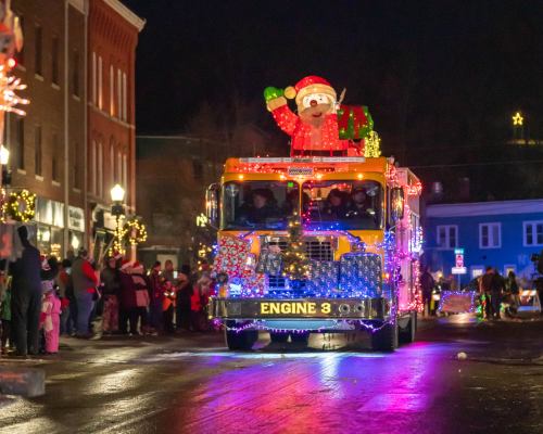 A festive fire truck decorated with lights and a Santa figure drives through a holiday parade at night.