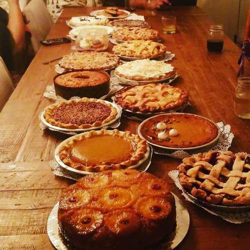 A long wooden table filled with various pies and desserts, including pumpkin, pecan, and fruit pies.