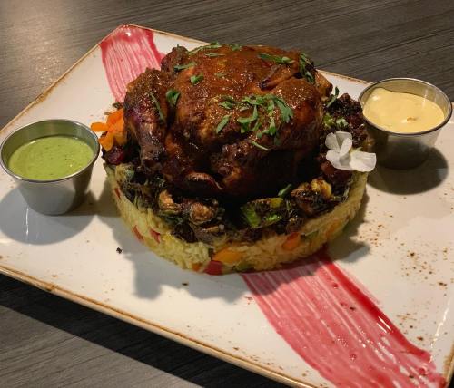 A beautifully plated dish featuring a whole roasted chicken on a bed of colorful rice, garnished with sauces and herbs.