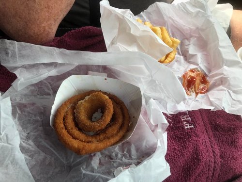 A close-up of a spiral onion ring in a paper container, surrounded by crumpled wrappers and a small portion of ketchup.